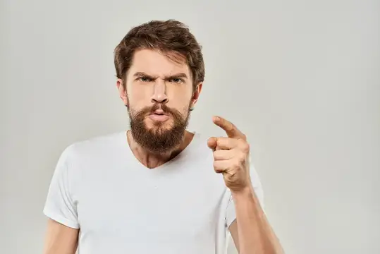 Бородатый мужчина в белой футболке строго грозит пальцем