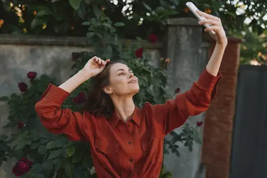 Молодая женщина в красной рубашке делает селфи на фоне розовых кустов
