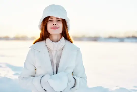 Рыжеволосая девушка в белой шапке и пальто улыбается на зимнем снегу