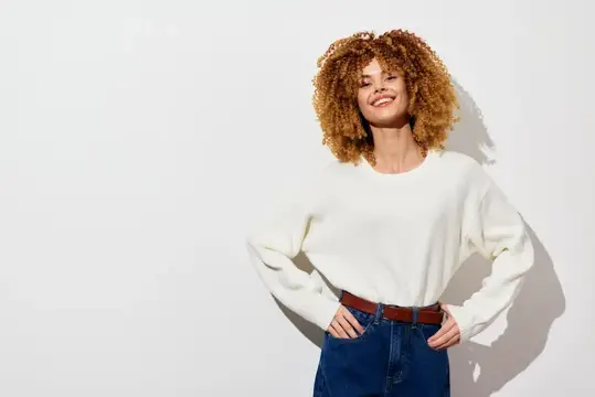 Улыбающаяся кудрявая девушка в белом свитере на светлом фоне