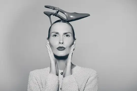 Светловолосая женщина с туфлей на голове держится за лицо
