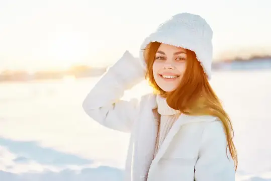 Рыжеволосая девушка в белой шапке улыбается на заснеженном поле
