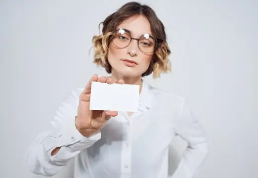 Женщина в очках и белой рубашке держит пустую визитку на светлом фоне