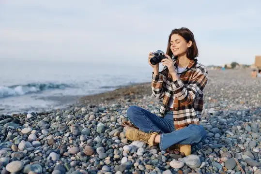 Молодая женщина с фотокамерой сидит на галечном пляже у моря