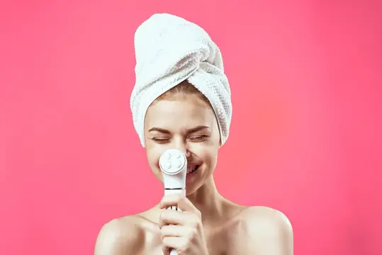 Улыбающаяся светловолосая девушка с полотенцем на голове и массажёром для лица на розовом фоне