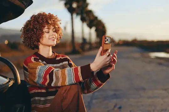 Кудрявая девушка в полосатом свитере фотографирует закат на телефон