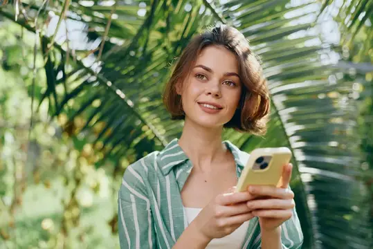 Улыбающаяся девушка с телефоном в руках на фоне пальмовых листьев