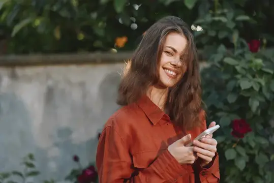 Улыбающаяся темноволосая девушка с телефоном в руках в саду
