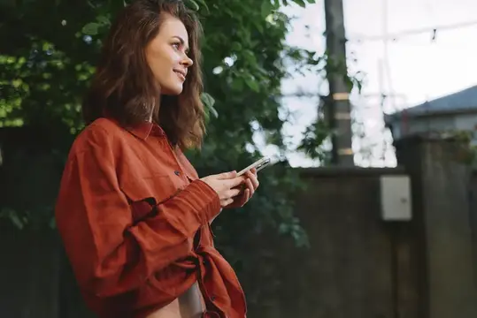 Улыбающаяся девушка в красной рубашке смотрит в телефон на улице