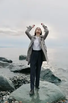 Молодая женщина в плаще стоит на камне у моря с поднятыми руками
