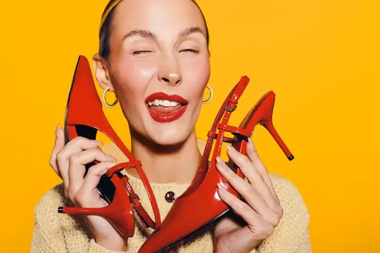 Улыбающаяся женщина с закрытыми глазами держит красные туфли на жёлтом фоне