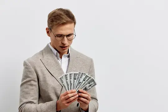 Бизнесмен в очках и бежевом пиджаке держит веер из долларовых купюр, глядя вниз, на фоне светло-серого фона