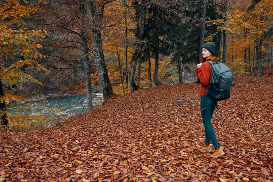 Рыжеволосая девушка с рюкзаком стоит в осеннем лесу у реки