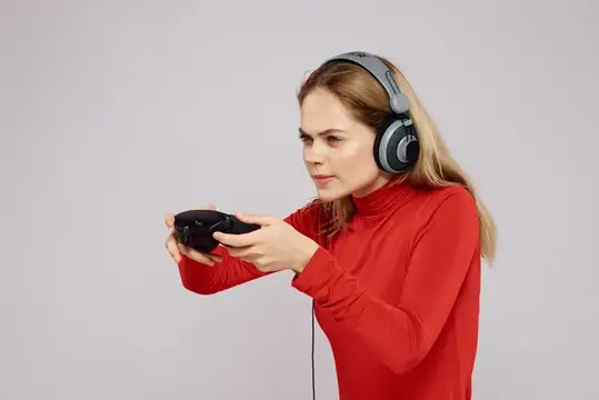 Женщина в наушниках с джойстиком в руках играет в игры, весёлый досуг, стиль жизни, развлечения, красная рубашка, серый фон