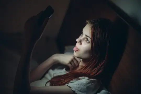 Рыжеволосая девушка лежит в темноте и смотрит в смартфон