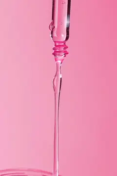 Капля косметической сыворотки стекает с пипетки на розовом фоне