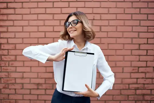 Улыбающаяся женщина в очках с планшетом на фоне кирпичной стены