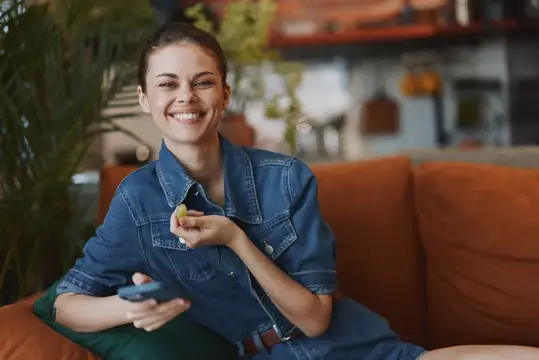 Улыбающаяся девушка в джинсовой куртке с телефоном на оранжевом диване