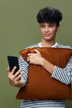 Кудрявый парень с подушкой смотрит в смартфон на зелёном фоне