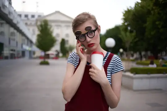 Рыжеволосая девушка в очках с кофе говорит по телефону на городской площади