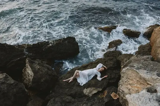 Девушка в белом платье лежит на камнях у бурного моря