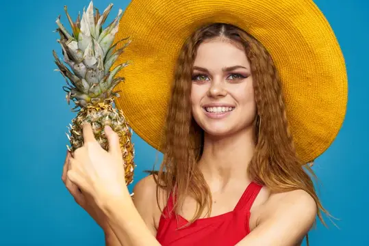 Улыбающаяся девушка в жёлтой шляпе держит ананас на синем фоне