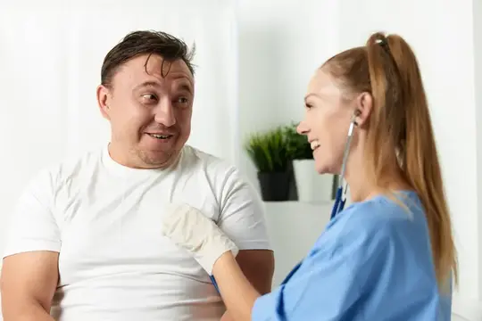 Врач с фонендоскопом осматривает улыбающегося пациента в белой футболке