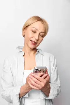 Улыбающаяся блондинка в белой рубашке смотрит в смартфон