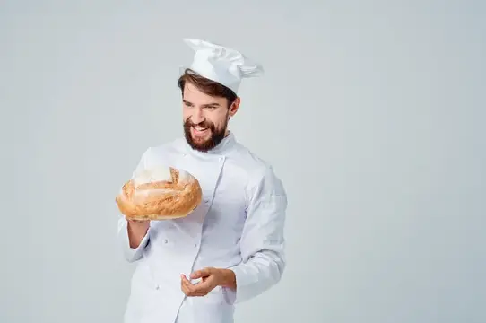 Улыбающийся бородатый пекарь в колпаке держит свежий каравай хлеба