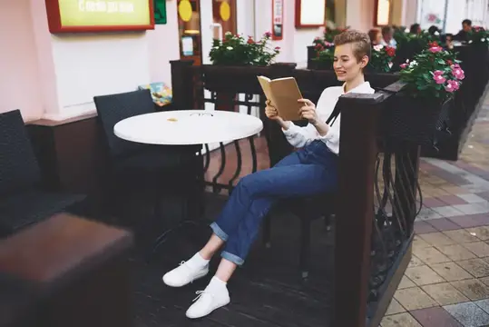 Девушка с короткой стрижкой читает книгу за столиком летнего кафе