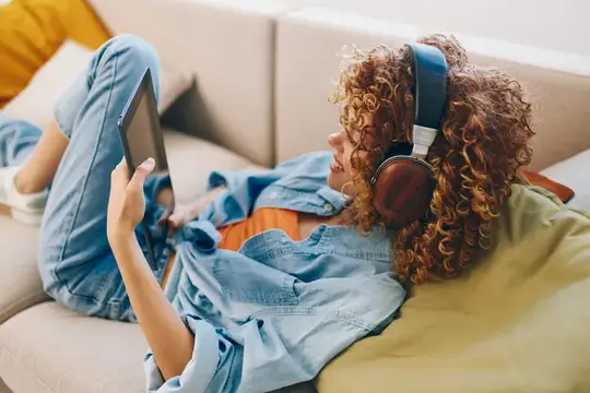 Кудрявая девушка в наушниках лежит на диване с планшетом