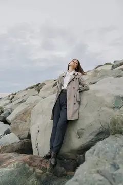 Расслабленная молодая женщина в пальто стоит на каменистом берегу с закрытыми глазами, наслаждаясь свежим воздухом