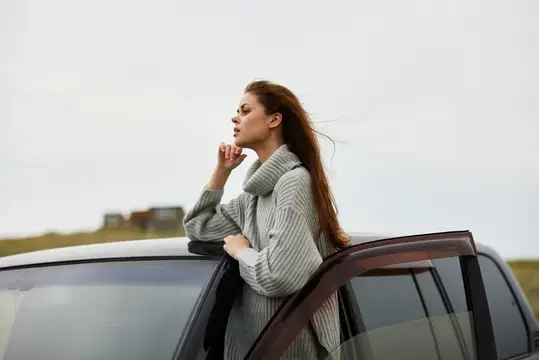 Молодая женщина в сером свитере стоит у открытой двери автомобиля