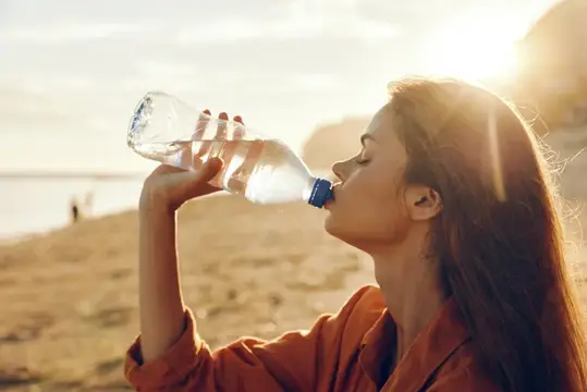 Молодая женщина пьёт воду из бутылки на закатном пляже
