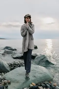 Молодая женщина в кепке и плаще стоит на камне у моря