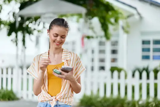 Улыбающаяся девушка в наушниках смотрит в смартфон на улице
