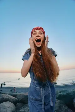 Молодая женщина в дениме и красной бандане выражает удивление и счастье на каменистом пляже на закате, сочетая современную бохо-вестерн моду с энергией образа жизни