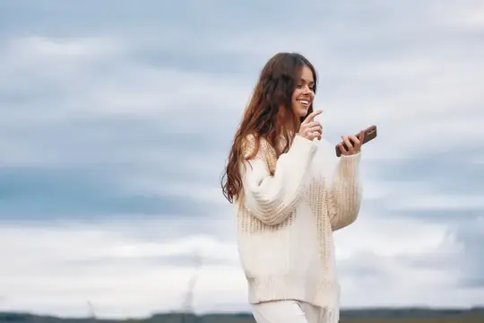 Улыбающаяся девушка в белом свитере смотрит на смартфон на фоне неба