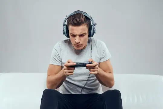 Мужчина в наушниках сидит на диване, мобильная игровая консоль, хобби, развлечения