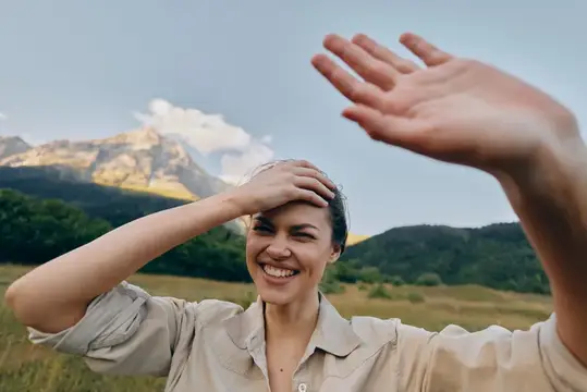 Улыбающаяся девушка закрывается рукой от солнца на фоне гор
