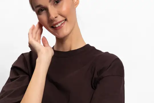 Улыбающаяся молодая женщина в коричневой футболке касается щеки рукой