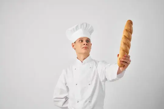 Повар в белом колпаке держит длинный багет на сером фоне
