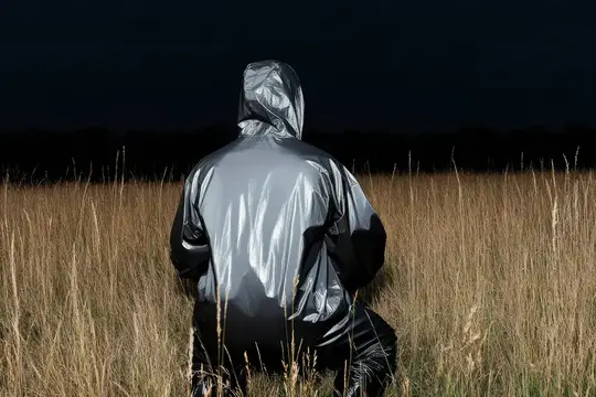 Человек в блестящем серебристом дождевике стоит спиной в сухом поле