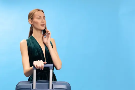 Привлекательная женщина в зеленом платье стоит с чемоданом и говорит по телефону с закрытыми глазами, изолированная на синем фоне