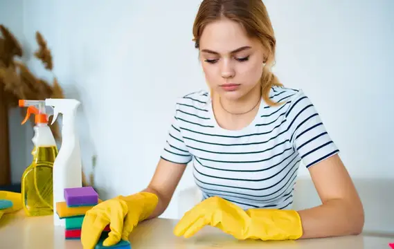 Молодая женщина в жёлтых перчатках отмывает стол с чистящими средствами