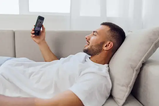 Молодой мужчина лежит на диване и смотрит в смартфон
