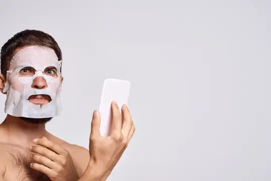 Мужчина с очищающей маской на лице рассматривает себя в зеркале на светлом фоне