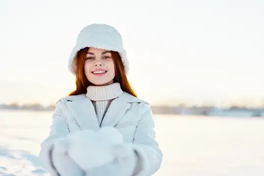 Улыбающаяся девушка в белой шапке и пуховике держит снег