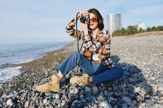 Улыбающаяся девушка в клетчатой рубашке фотографирует на камеру у моря