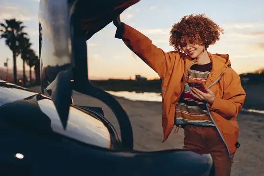 Кудрявая девушка с телефоном у открытого капота автомобиля на закате